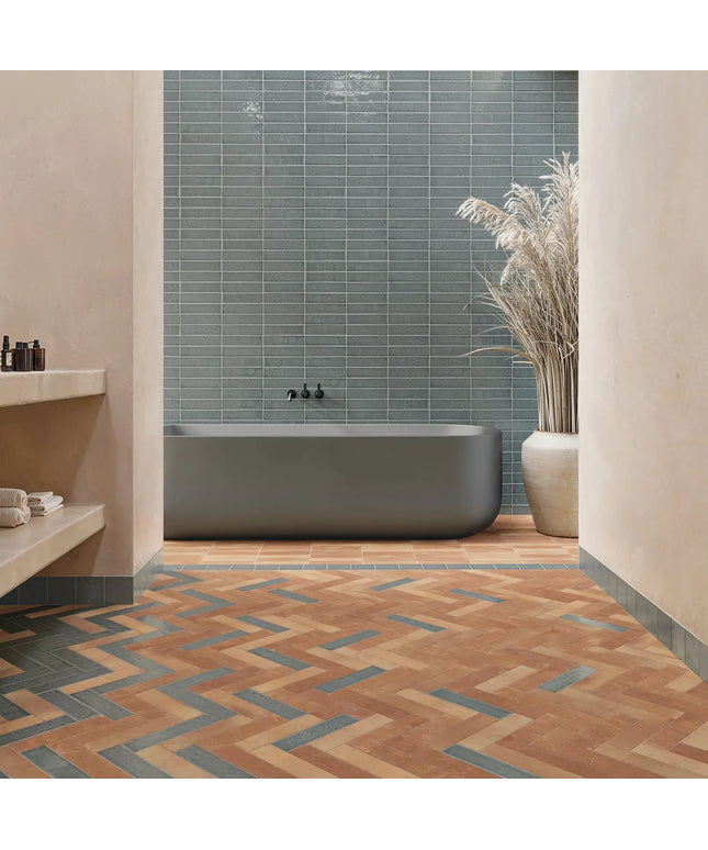 A modern bathroom interior with a freestanding matte gray bathtub, blue-gray rectangular wall tiles, terracotta and blue chevron floor tiles, beige walls, a built-in shelf, and a tall ceramic vase with dried grass.