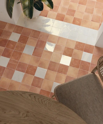 Terracotta and cream checkered floor tiles shown in a room setting with a wooden table, a woven chair, and a potted plant.