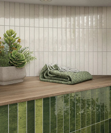 A styled interior scene showing a countertop with a folded green quilted blanket, a potted succulent, and a backsplash made of glossy vertical green tiles against a light beige tiled wall.