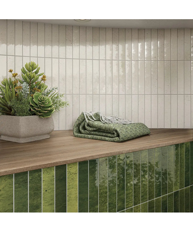 A styled interior scene showing a countertop with a folded green quilted blanket, a potted succulent, and a backsplash made of glossy vertical green tiles against a light beige tiled wall.