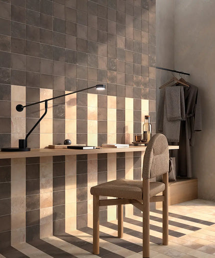 A modern interior scene featuring a wall of square tiles in dark gray and warm beige tones, with a wooden desk, black adjustable desk lamp, upholstered wooden chair, and a hanging robe.