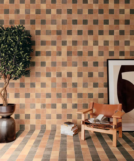 A styled interior scene featuring a checkered tile wall and striped tile floor in warm terracotta, beige, and dark gray tones, with a potted tree, a wooden chair, books, and a framed abstract print.