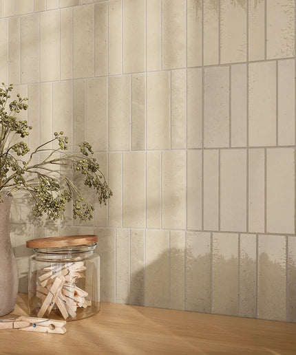 Beige rectangular subway wall tiles with a subtle distressed texture, styled on a light wood countertop next to a vase with dried greenery and a glass jar.