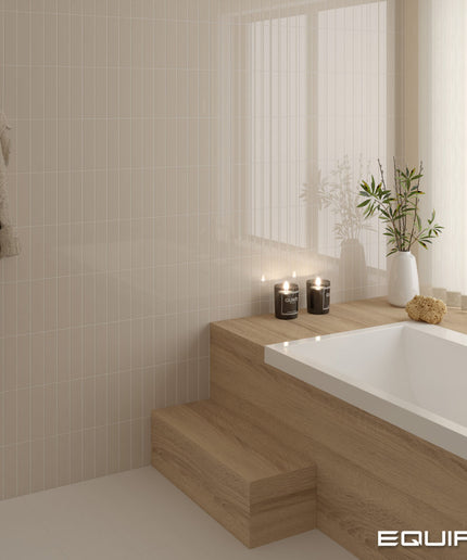 A modern bathroom corner with a white bathtub set into light wood steps, beige tiled walls, a hanging robe, candles, and a white vase with greenery. The word EQUIPE appears in the bottom right corner.