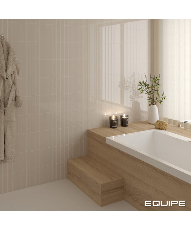 A modern bathroom corner with a white bathtub set into light wood steps, beige tiled walls, a hanging robe, candles, and a white vase with greenery. The word EQUIPE appears in the bottom right corner.