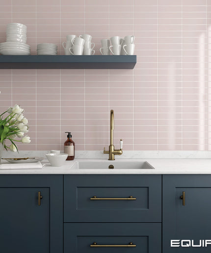 A modern kitchen scene with navy blue cabinets, a white countertop, a gold faucet, and a pale pink tiled backsplash. A floating shelf holds white dishes, and a vase of tulips sits on the counter.