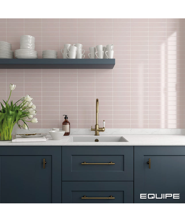 A modern kitchen scene with navy blue cabinets, a white countertop, a gold faucet, and a pale pink tiled backsplash. A floating shelf holds white dishes, and a vase of tulips sits on the counter.