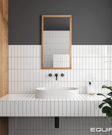 Modern bathroom vanity setup with a white rectangular vessel sink, white subway tile backsplash, black faucet hardware, a rectangular wood-framed mirror, and a dark charcoal wall. A green plant is visible on the right, and the word EQUIPE appears in the bottom right corner.