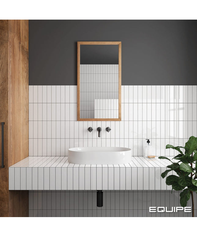 Modern bathroom vanity setup with a white rectangular vessel sink, white subway tile backsplash, black faucet hardware, a rectangular wood-framed mirror, and a dark charcoal wall. A green plant is visible on the right, and the word EQUIPE appears in the bottom right corner.