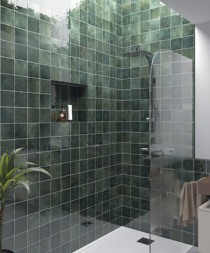 A modern bathroom corner featuring dark green glossy square wall tiles, a glass shower door, a built-in niche shelf, and a small potted plant.