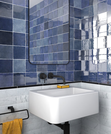 A modern bathroom corner featuring glossy blue square wall tiles, a white floating sink, a black faucet, and a yellow towel on a black rack.