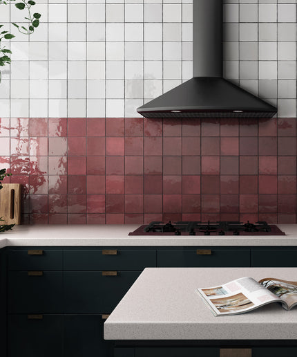 A modern kitchen interior with glossy square tiles in white on the upper wall and dusty rose pink on the lower backsplash. A black range hood hangs above a gas cooktop, with dark green cabinetry below and a light stone countertop in the foreground.