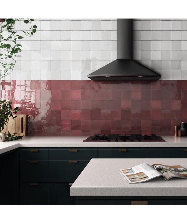 A modern kitchen interior with glossy square tiles in white on the upper wall and dusty rose pink on the lower backsplash. A black range hood hangs above a gas cooktop, with dark green cabinetry below and a light stone countertop in the foreground.