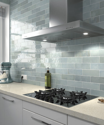 A modern kitchen scene featuring glossy light blue-gray subway tiles on the backsplash, a stainless steel range hood, a black gas cooktop, and a light stone countertop with a stand mixer and other kitchen items.