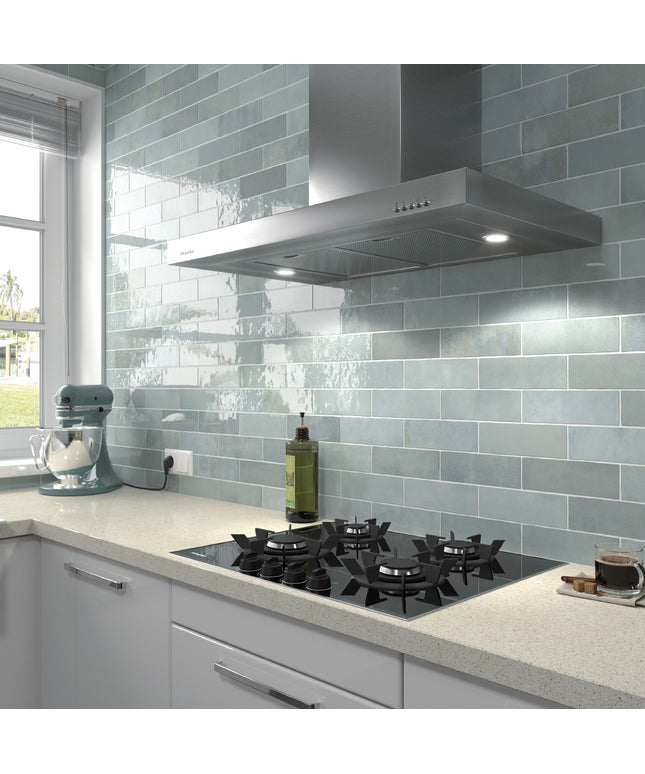 A modern kitchen scene featuring glossy light blue-gray subway tiles on the backsplash, a stainless steel range hood, a black gas cooktop, and a light stone countertop with a stand mixer and other kitchen items.