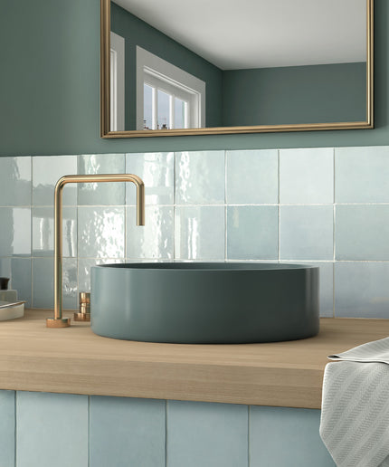 A modern bathroom scene featuring a matte slate blue vessel sink paired with a brushed brass faucet, set on a light wood countertop against a blue tiled backsplash and a matching slate blue wall with a gold-framed mirror.