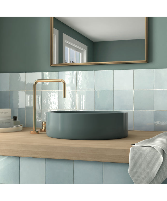 A modern bathroom scene featuring a matte slate blue vessel sink paired with a brushed brass faucet, set on a light wood countertop against a blue tiled backsplash and a matching slate blue wall with a gold-framed mirror.