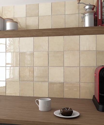 Beige glossy ceramic wall tiles installed on a kitchen backsplash above a wooden countertop, with a white mug, a small plate with a pastry, and a red coffee machine visible.