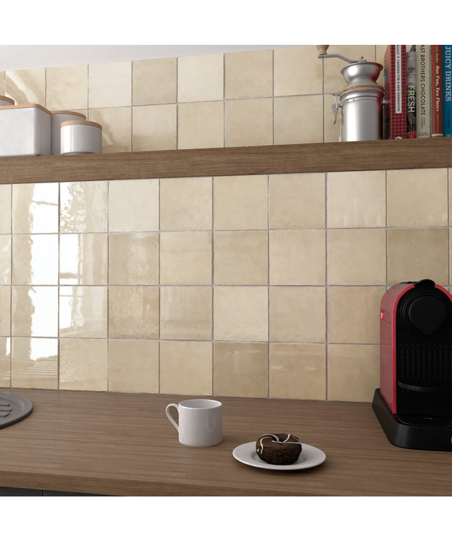 Beige glossy ceramic wall tiles installed on a kitchen backsplash above a wooden countertop, with a white mug, a small plate with a pastry, and a red coffee machine visible.