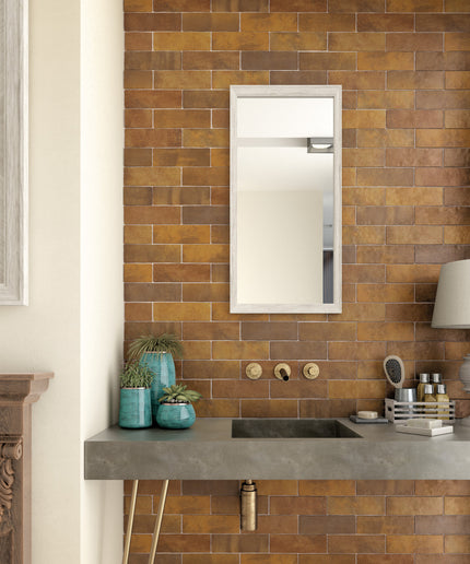 A modern bathroom vanity setup featuring a rectangular wall mirror with a white frame, set against a warm brown and gold subway tile wall. Below the mirror is a gray stone countertop with a built-in sink, gold wall-mounted fixtures, and decorative teal vases.