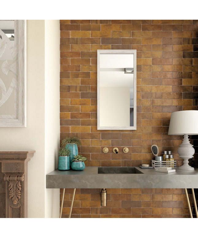 A modern bathroom vanity setup featuring a rectangular wall mirror with a white frame, set against a warm brown and gold subway tile wall. Below the mirror is a gray stone countertop with a built-in sink, gold wall-mounted fixtures, and decorative teal vases.