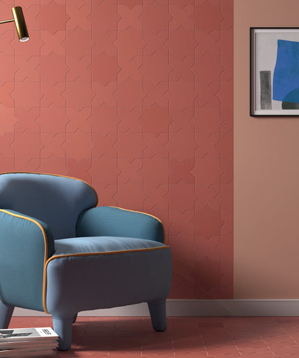 A styled interior scene showing a blue upholstered armchair with gold piping against a terracotta and peach wall, with a black floor lamp, framed abstract art, and a tiled floor.