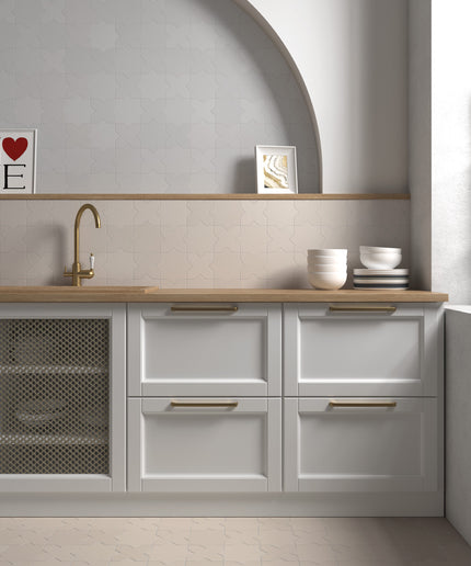 A modern kitchen cabinet unit with white shaker-style drawers and a wood-look countertop, featuring a brass gooseneck faucet, a mesh-front lower cabinet, and gold-toned drawer pulls. The scene is styled with ceramic bowls, a small bottle, and framed art against a light gray wall with a built-in shelf and arched niche.