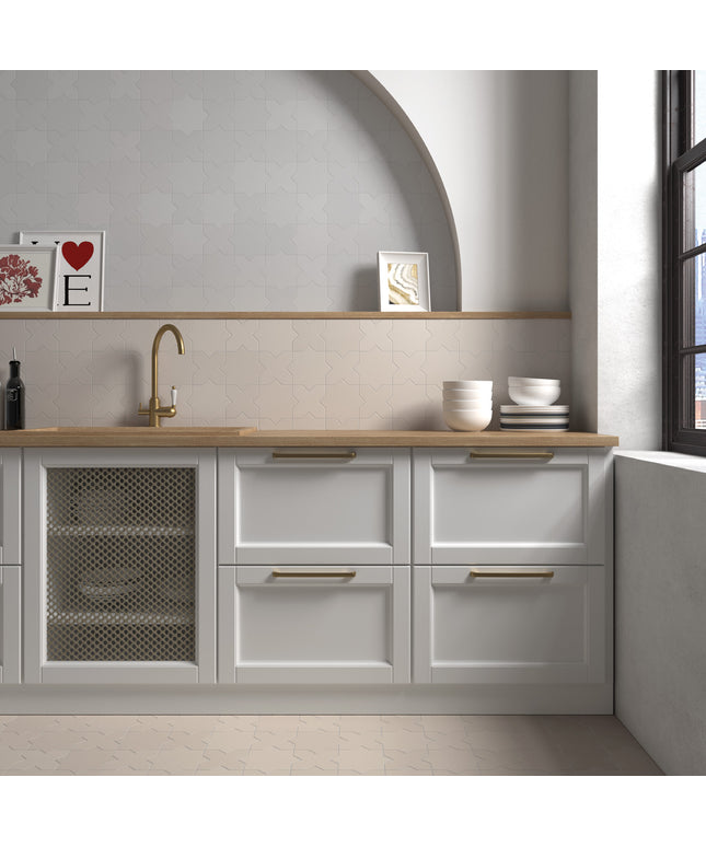 A modern kitchen cabinet unit with white shaker-style drawers and a wood-look countertop, featuring a brass gooseneck faucet, a mesh-front lower cabinet, and gold-toned drawer pulls. The scene is styled with ceramic bowls, a small bottle, and framed art against a light gray wall with a built-in shelf and arched niche.