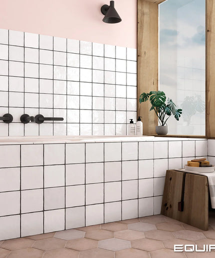 A modern bathroom interior with white square wall tiles, a bathtub, black faucet hardware, a pink wall, a wooden window frame, a potted plant, and a small wooden stool with a towel. The word EQUIPE appears in the bottom right corner.