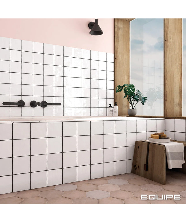 A modern bathroom interior with white square wall tiles, a bathtub, black faucet hardware, a pink wall, a wooden window frame, a potted plant, and a small wooden stool with a towel. The word EQUIPE appears in the bottom right corner.