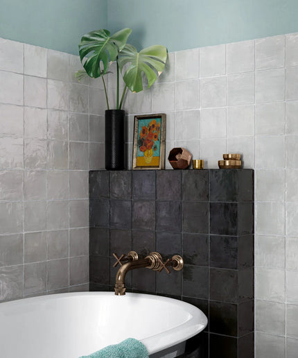 A styled bathroom corner featuring a white bathtub with a matte bronze faucet, set against a light gray tiled wall and a dark charcoal tiled accent wall. A black vase with a green monstera plant and a small colorful framed print sit on top of the dark tiled wall, alongside a teal towel draped over the tub edge.