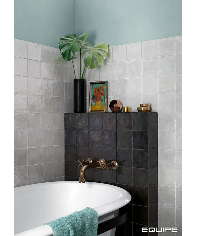 A styled bathroom corner featuring a white bathtub with a matte bronze faucet, set against a light gray tiled wall and a dark charcoal tiled accent wall. A black vase with a green monstera plant and a small colorful framed print sit on top of the dark tiled wall, alongside a teal towel draped over the tub edge.