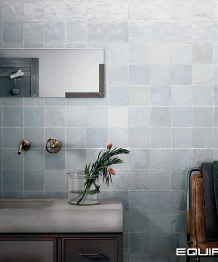 A bathroom wall covered in square light blue-gray tiles, with a concrete-look sink countertop, brass faucet hardware, a mirror, a vase with flowers, and towels on a rack. The word EQUIPE appears in the bottom right corner.
