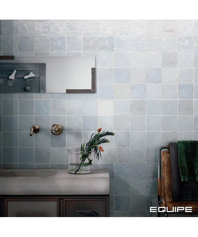 A bathroom wall covered in square light blue-gray tiles, with a concrete-look sink countertop, brass faucet hardware, a mirror, a vase with flowers, and towels on a rack. The word EQUIPE appears in the bottom right corner.