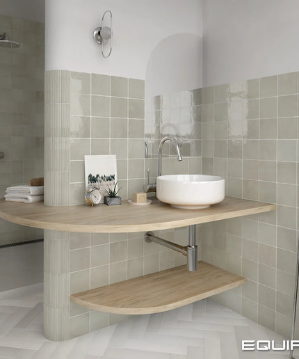 A modern floating bathroom vanity with a curved light wood top and lower shelf, a white vessel sink, and a chrome faucet, set against beige tiled walls.