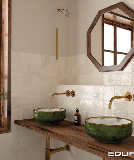 A styled bathroom vanity featuring two green ceramic vessel sinks with gold trim on a rustic wooden countertop, paired with gold wall-mounted faucets and a hexagonal wood-framed mirror on a tiled wall.