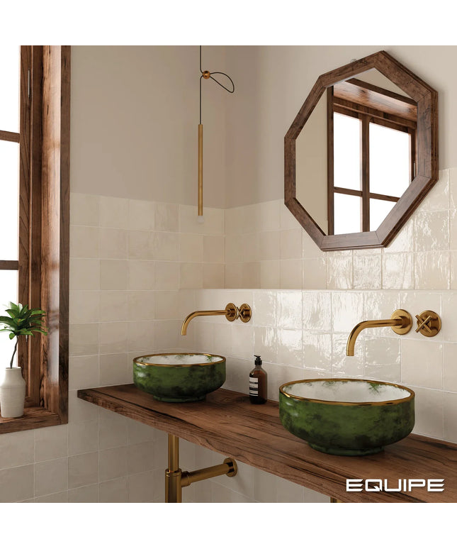 A styled bathroom vanity featuring two green ceramic vessel sinks with gold trim on a rustic wooden countertop, paired with gold wall-mounted faucets and a hexagonal wood-framed mirror on a tiled wall.