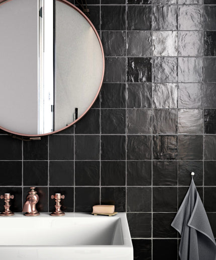 Black glossy square wall tiles in a modern bathroom setting, featuring a white sink, copper faucet, a round mirror, and a gray towel. The word 'EQUIPE' is visible in the bottom right corner.