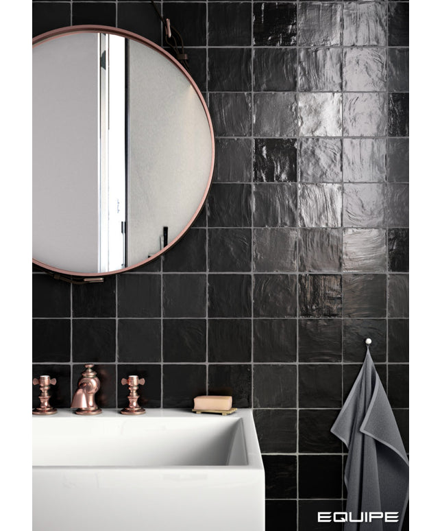 Black glossy square wall tiles in a modern bathroom setting, featuring a white sink, copper faucet, a round mirror, and a gray towel. The word 'EQUIPE' is visible in the bottom right corner.