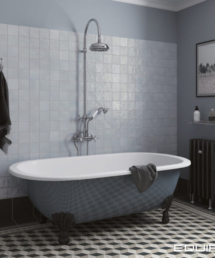 A freestanding clawfoot bathtub in a blue-gray finish with a white interior, set in a styled bathroom with light blue tiled walls, a chrome shower fixture, and a black patterned floor.