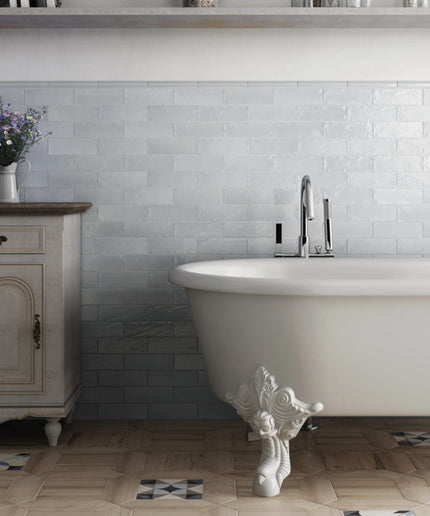 A white freestanding clawfoot bathtub in a styled bathroom setting with a light gray subway tile backsplash, light wood floor, and a vintage-style cabinet on the left.