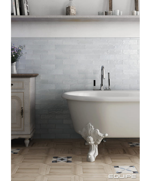 A white freestanding clawfoot bathtub in a styled bathroom setting with a light gray subway tile backsplash, light wood floor, and a vintage-style cabinet on the left.