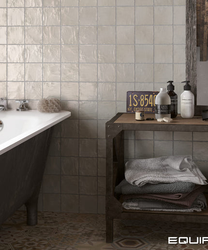 A styled bathroom scene with a white clawfoot tub on the left, a dark wood shelf with towels and toiletries on the right, and textured beige wall tiles behind. The word EQUIPE appears in the bottom right corner.