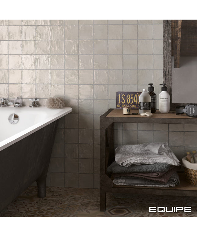 A styled bathroom scene with a white clawfoot tub on the left, a dark wood shelf with towels and toiletries on the right, and textured beige wall tiles behind. The word EQUIPE appears in the bottom right corner.