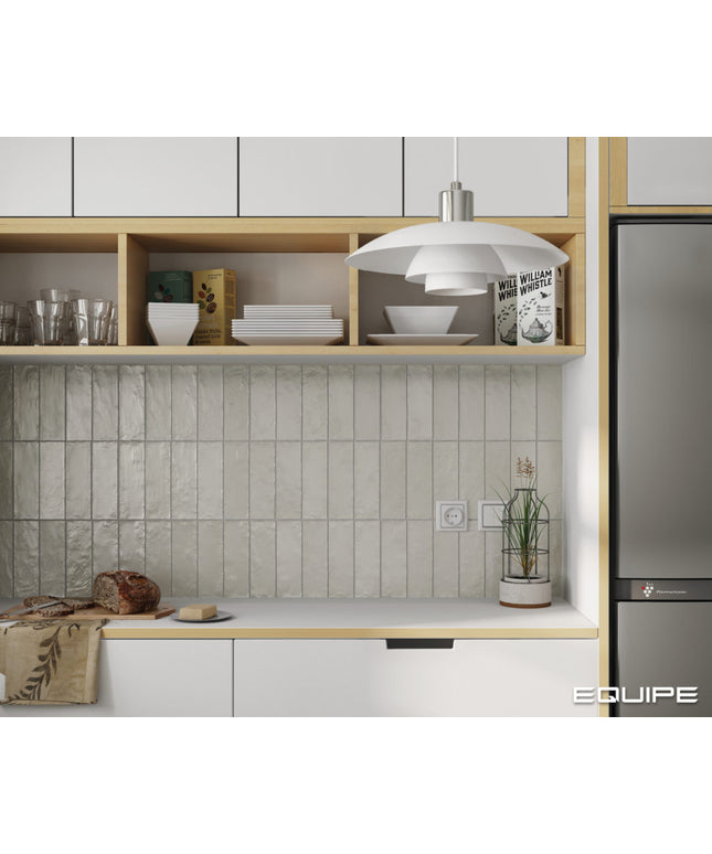 A modern kitchen interior featuring a textured light gray tile backsplash, light wood open shelving with white dishes, a white pendant light, and a stainless steel refrigerator. The word 'EQUIPE' appears in the bottom right corner.
