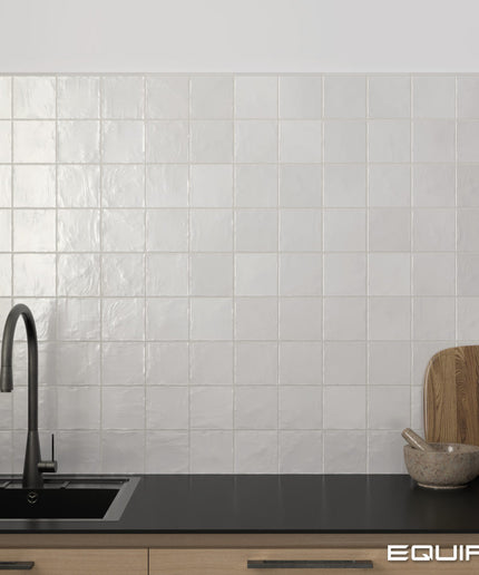 White square glossy ceramic wall tiles installed on a kitchen backsplash behind a black countertop with a dark faucet, wooden cutting board, and small bowl.