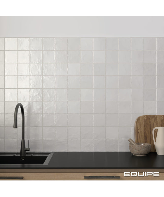 White square glossy ceramic wall tiles installed on a kitchen backsplash behind a black countertop with a dark faucet, wooden cutting board, and small bowl.