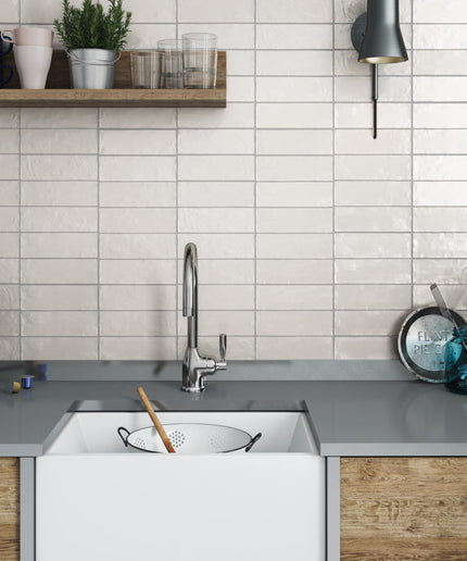 A modern kitchen scene featuring a white farmhouse-style undermount sink with a chrome faucet, set against a white subway tile backsplash and a gray countertop. A wooden floating shelf with glassware and a potted plant is mounted above, and a black wall sconce is on the right.