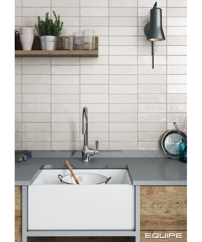 A modern kitchen scene featuring a white farmhouse-style undermount sink with a chrome faucet, set against a white subway tile backsplash and a gray countertop. A wooden floating shelf with glassware and a potted plant is mounted above, and a black wall sconce is on the right.