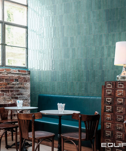 Restaurant interior with teal tiled walls, wooden chairs, and a lamp.